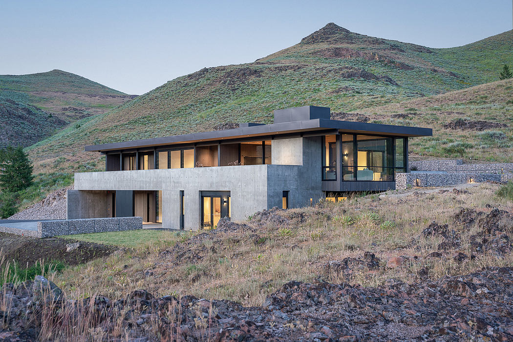 A modern, two-story house with large windows, a flat roof, and a stone wall facade nestled in a mountainous landscape.