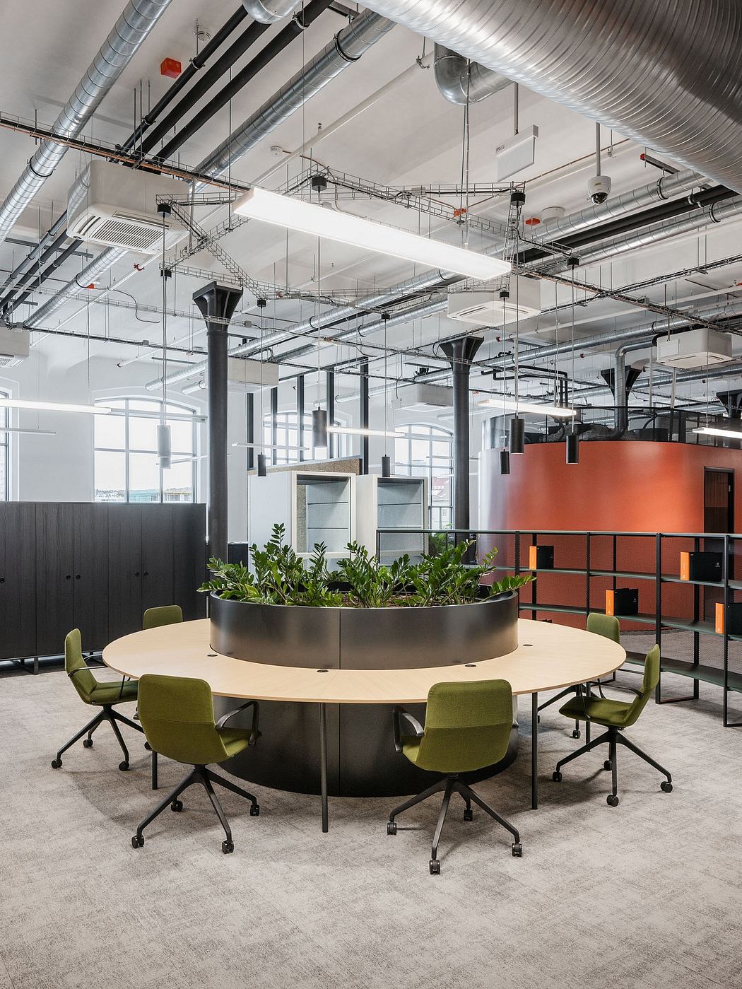 A modern, open-concept office space with exposed ducts, green chairs, and a planter centerpiece.