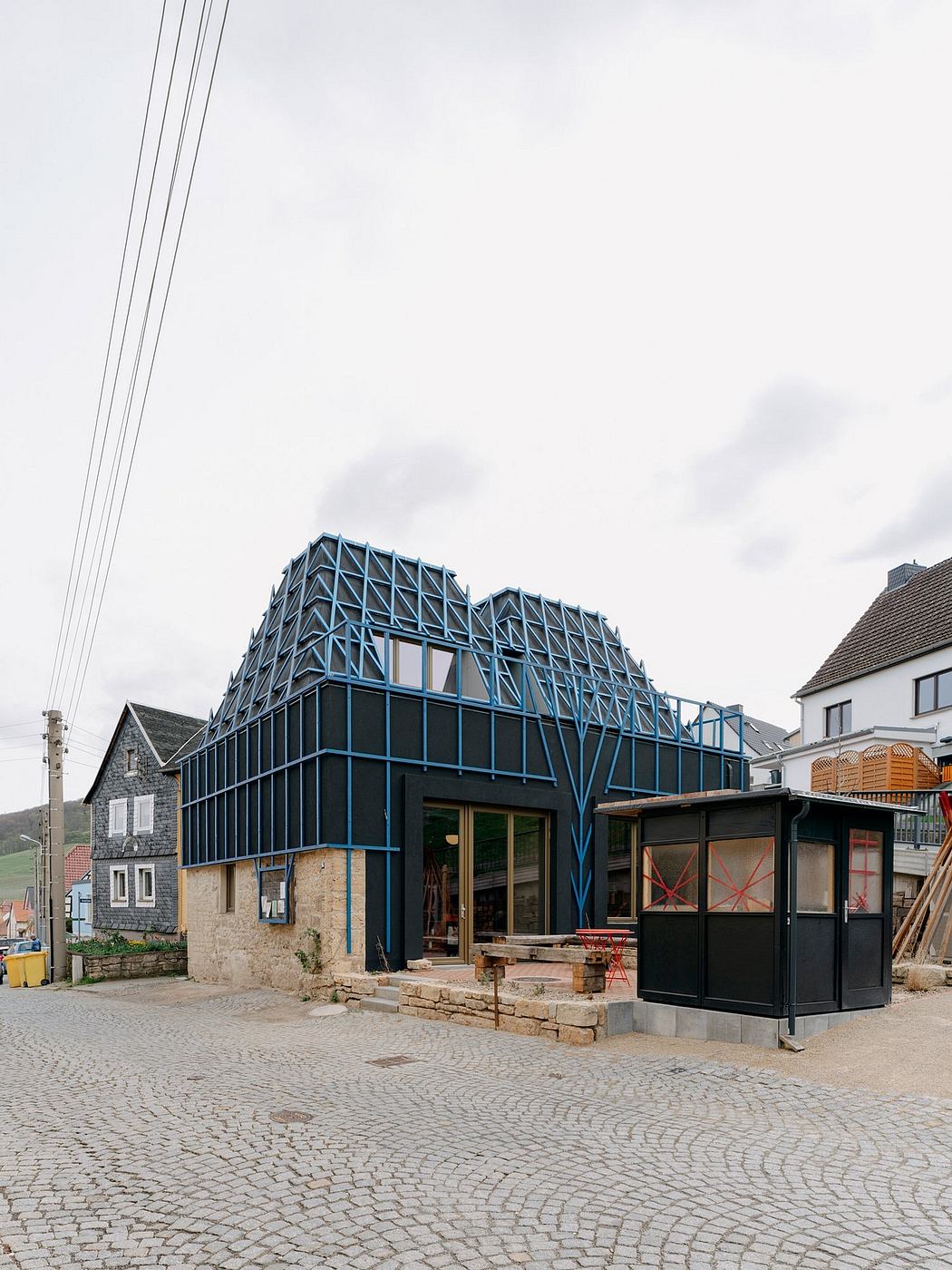 A modern, triangular structure with a distinctive blue metal framework stands in a village setting.