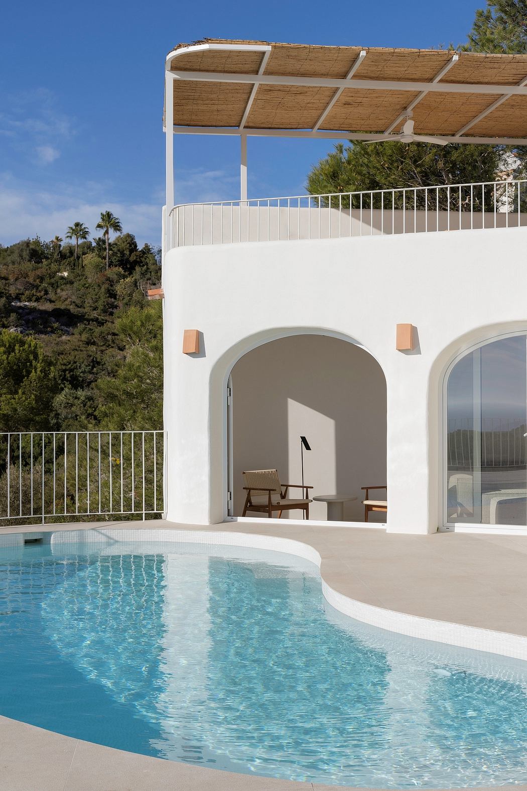 A modern white villa with arched entryways, a thatched roof, and a swimming pool overlooking lush greenery.