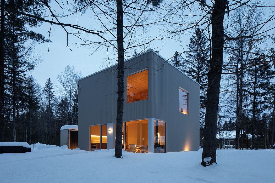 Modern, minimalist cabin in a snowy forest with warm lighting and large windows.