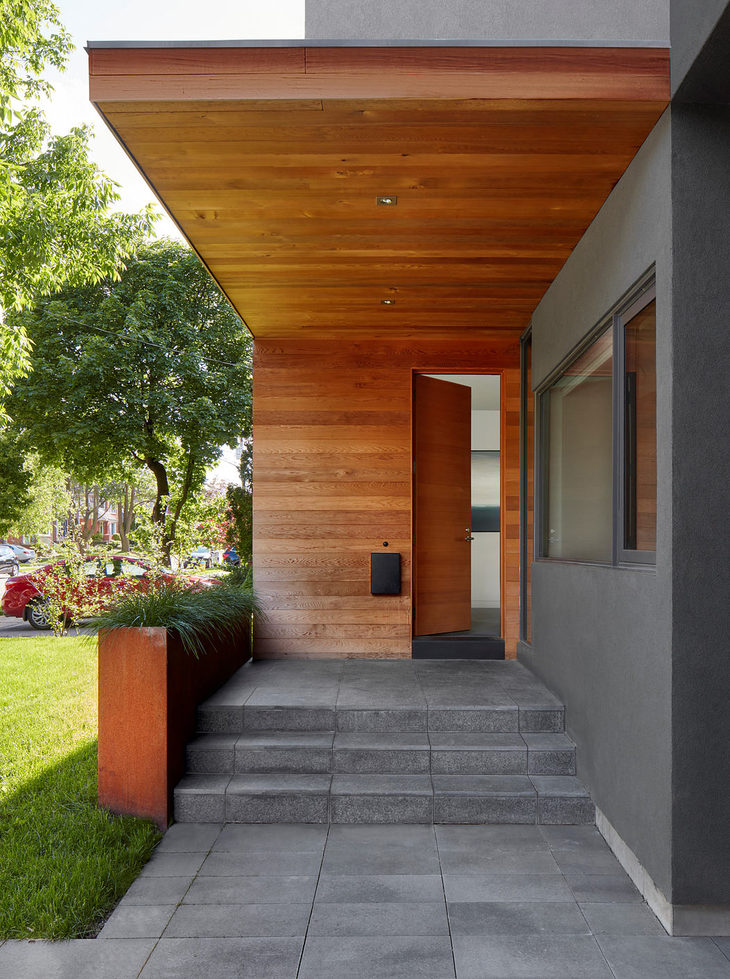Wooden overhang, gray stone steps, modern facade with glass door, lush greenery.