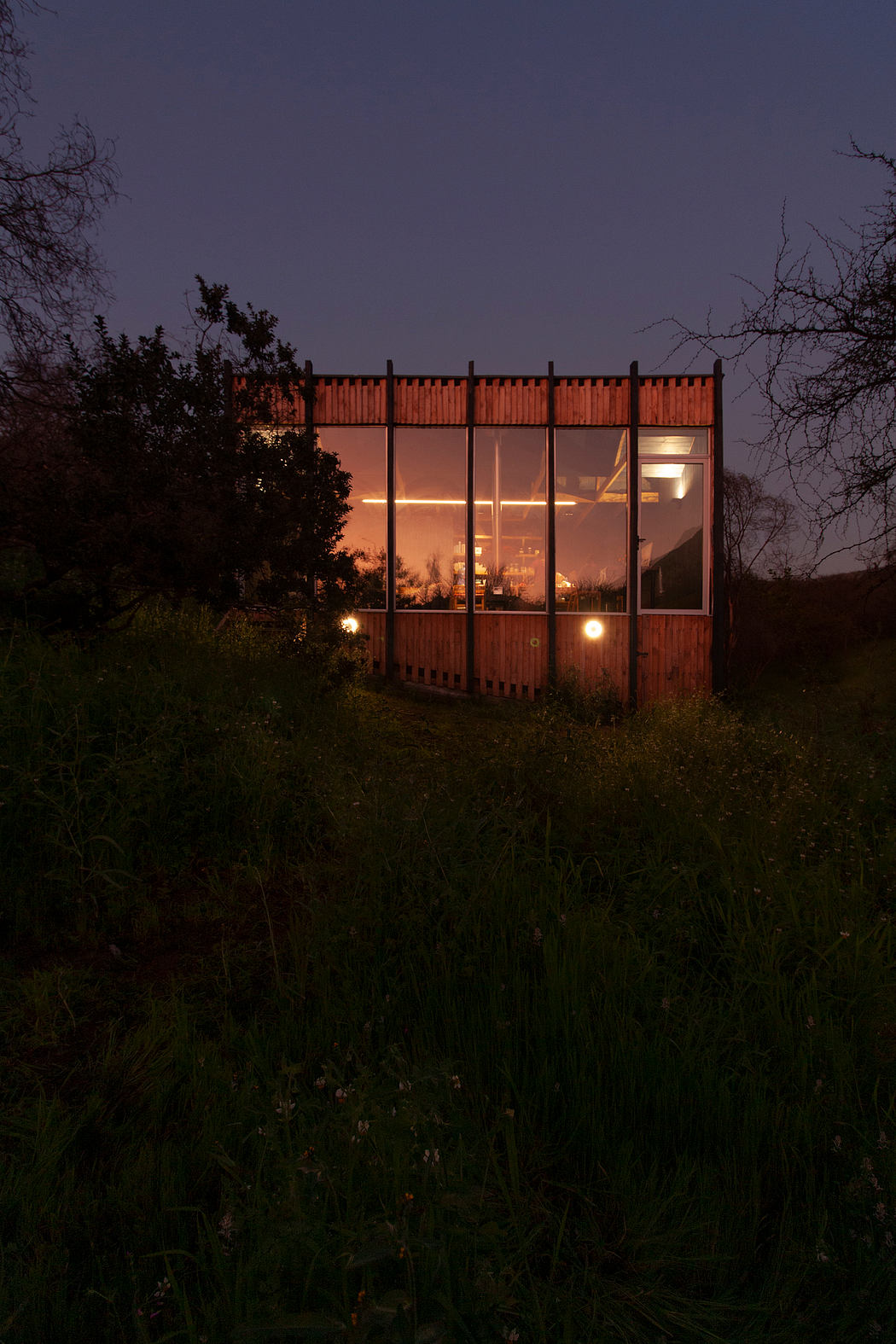 A modern, glass-enclosed structure with wooden paneled exterior stands in a grassy field.