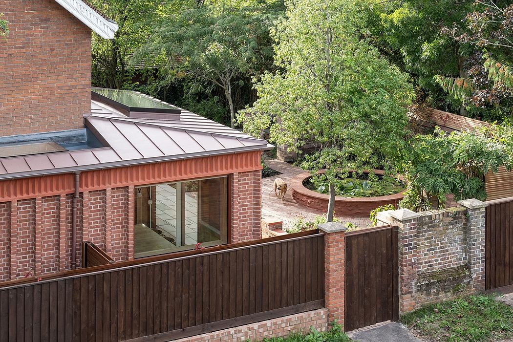Brick building with a slanted metal roof, surrounded by lush greenery and a wooden fence.