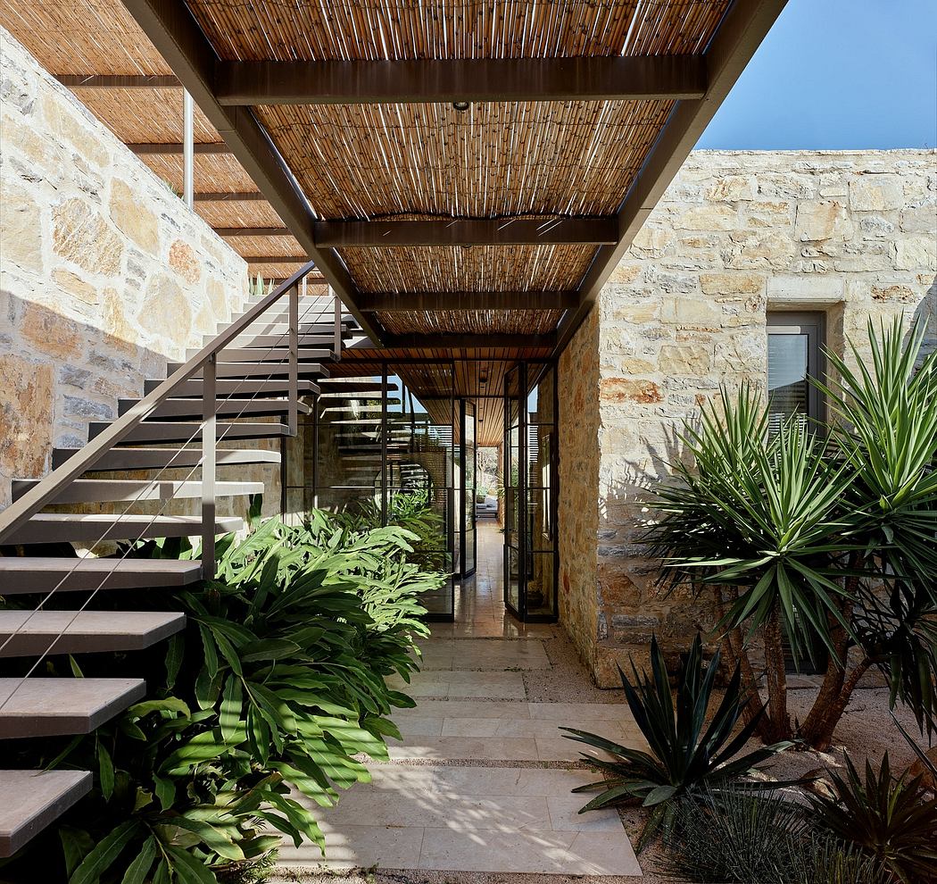 A rustic stone hallway with a thatched roof, wooden stairs, and lush tropical plants.