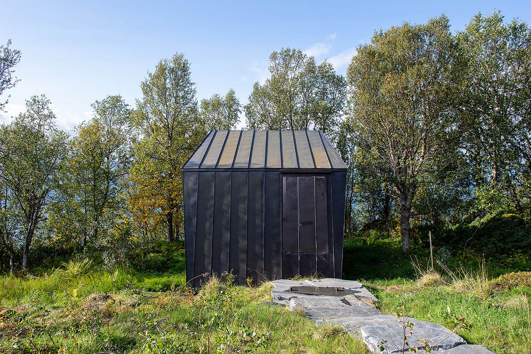 A small, angular, black steel structure with a pitched metal roof amid lush greenery.