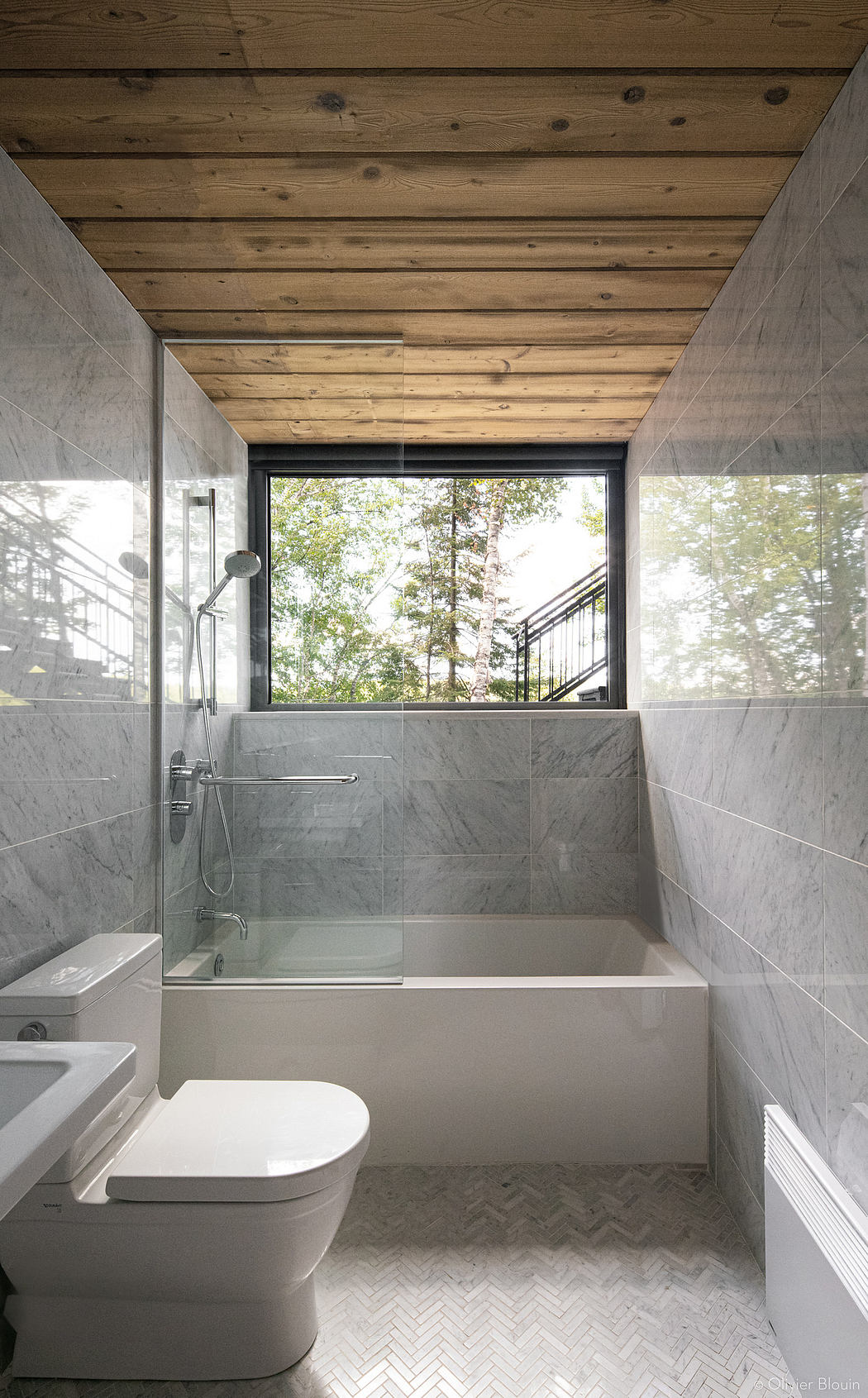 Modern bathroom with wood-paneled ceiling, marble tiles, and large window overlooking nature.