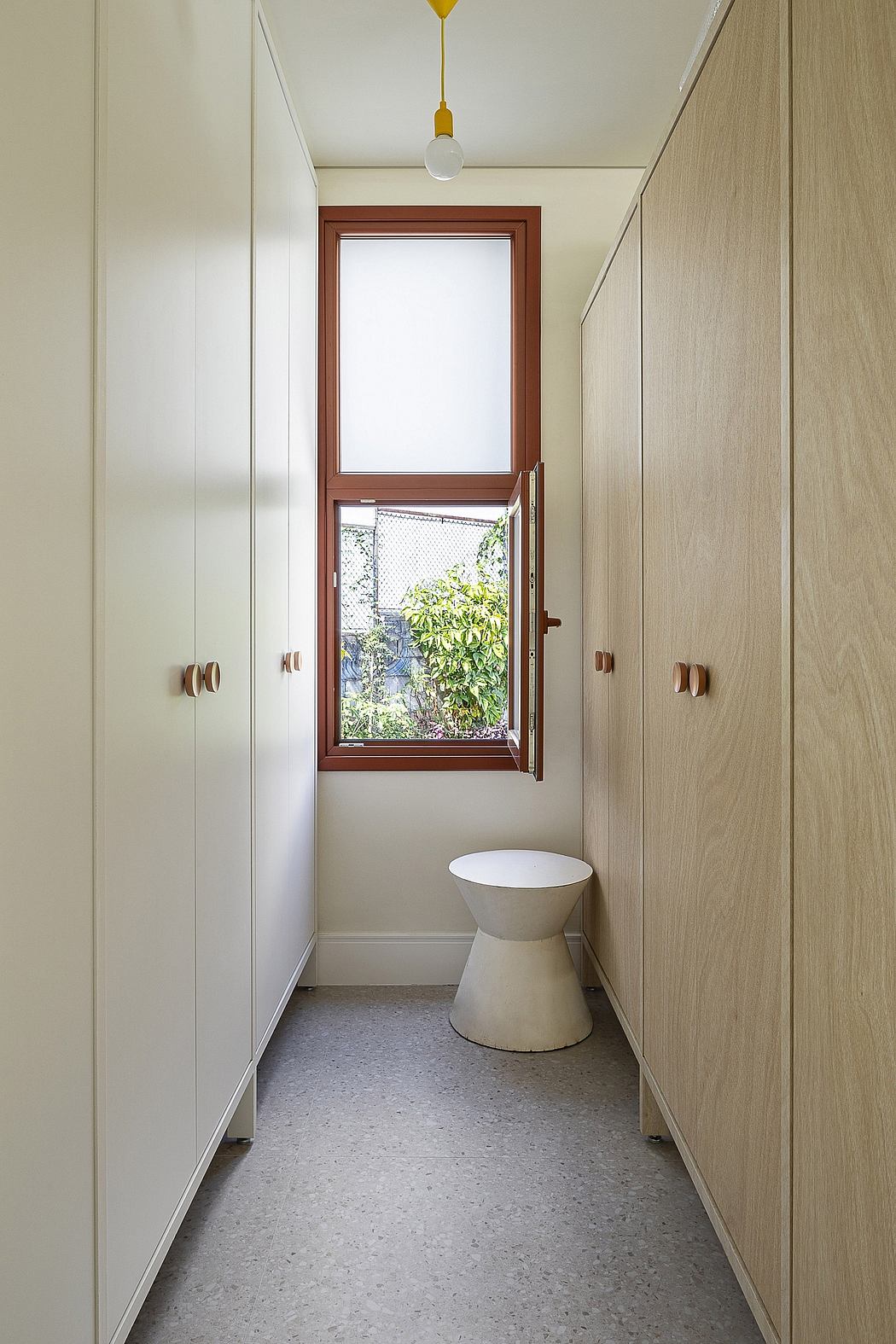 A narrow hallway featuring built-in wooden cabinets, a window with nature views, and a pedestal sink.