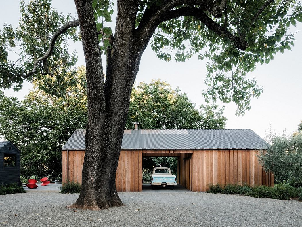 A wooden barn-style structure with a car parked underneath a large, overarching tree.