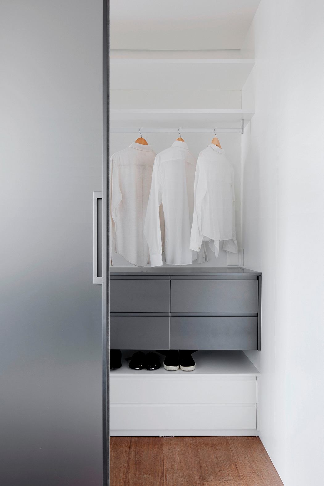 Clean, minimalist closet with wood floor, white shirts, and drawers in a neutral gray color.