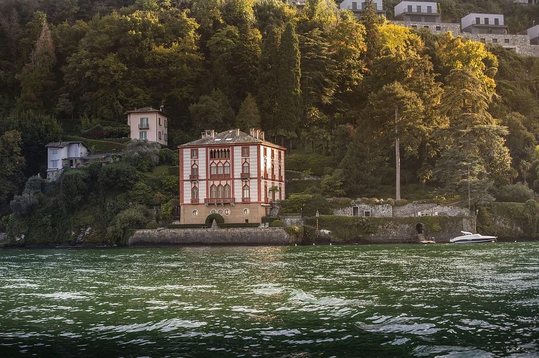 A historic, ornate red building stands on a lush, forested shoreline beside a boat-dotted lake.