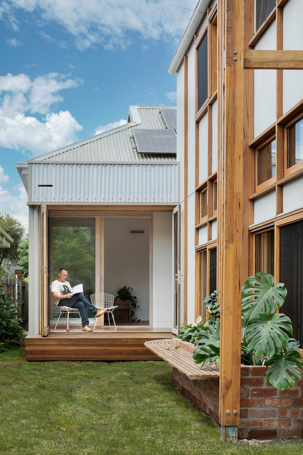 Rustic modern home with wooden and metal accents, lush greenery, and a person reading on the porch.