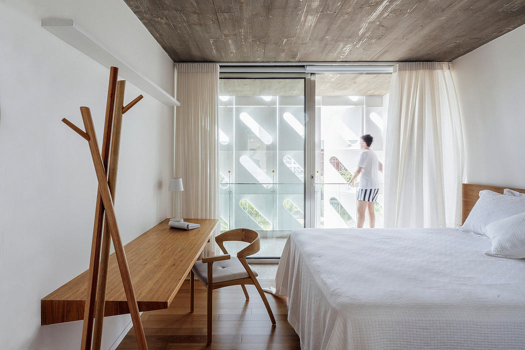 A modern, minimalist bedroom with a wooden desk, chair, and large windows overlooking nature.