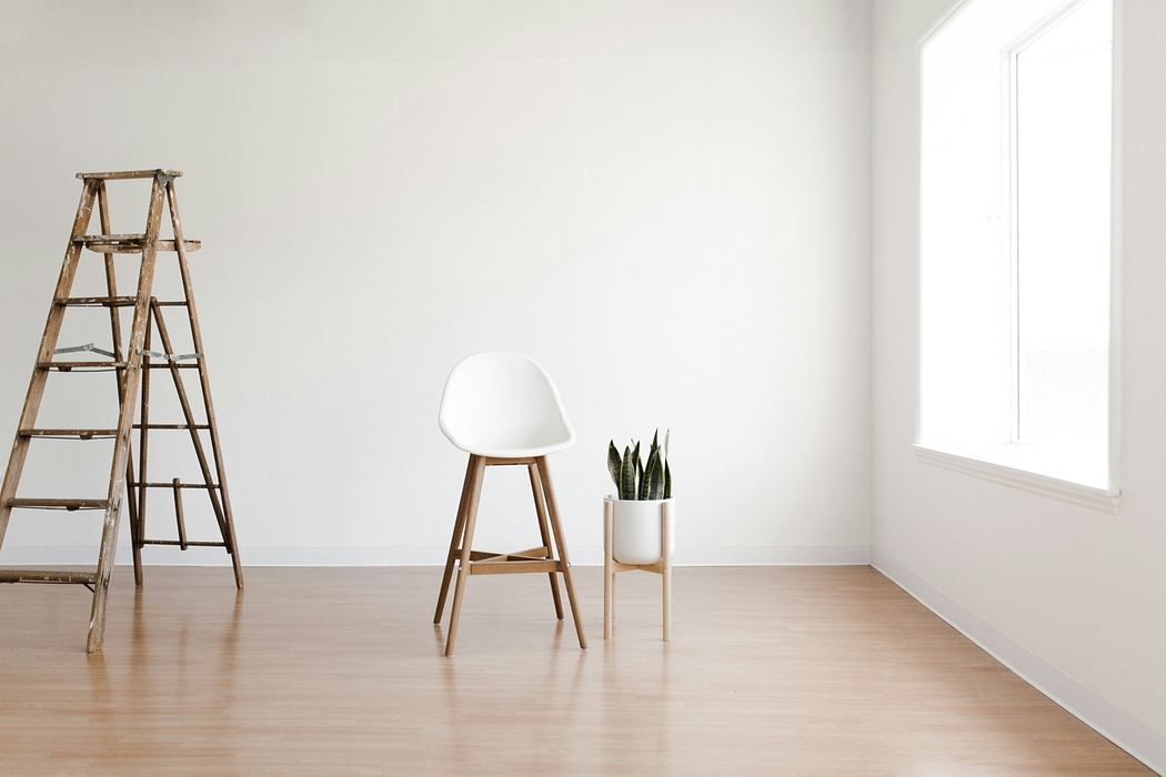 Minimalist room with white walls, wooden floor, ladder, and modern white chair with indoor plant.