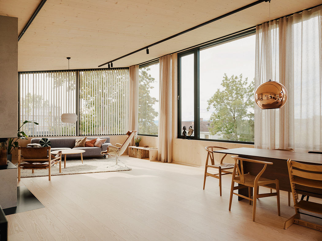 A cozy, modern living room with warm wooden furniture, large windows, and a copper pendant lamp.
