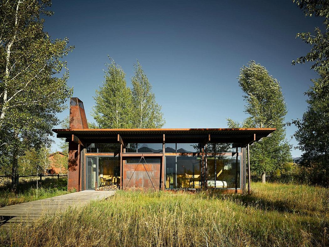 The image depicts a modern, glass-walled structure with a wooden exterior and an elevated porch, surrounded by lush greenery.