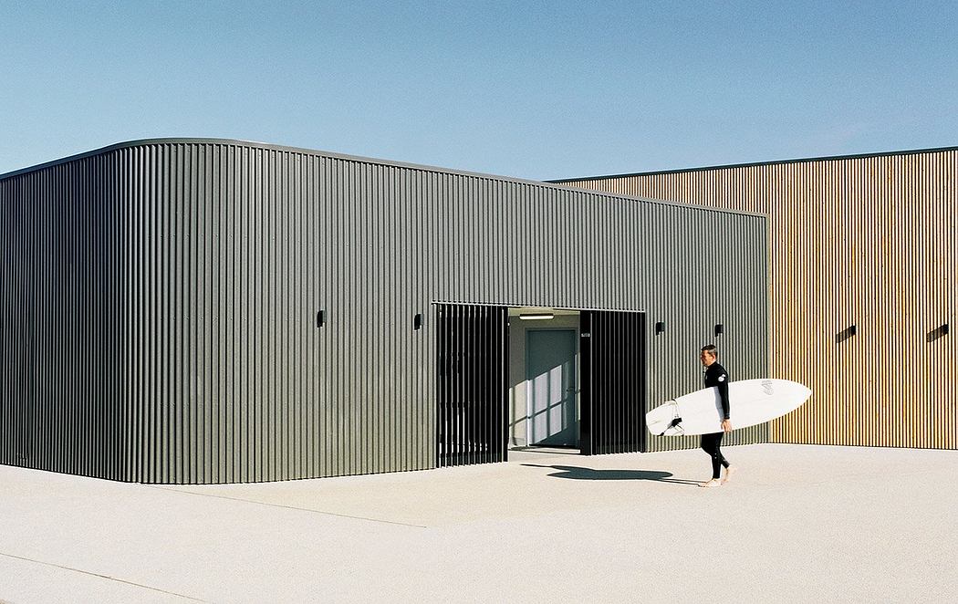 A rectangular, black metal building with a surfboard-toting person entering through the large doorway.