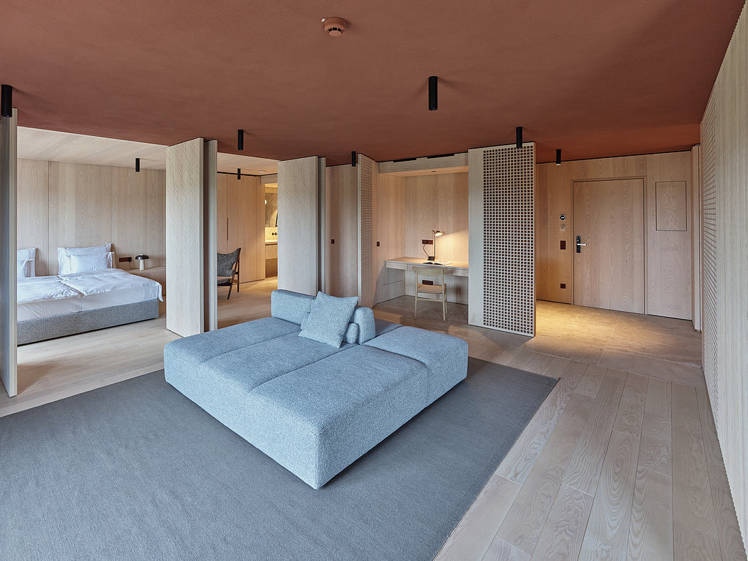 A modern, minimalist hotel suite with wood paneling, gray sectional, and recessed lighting.