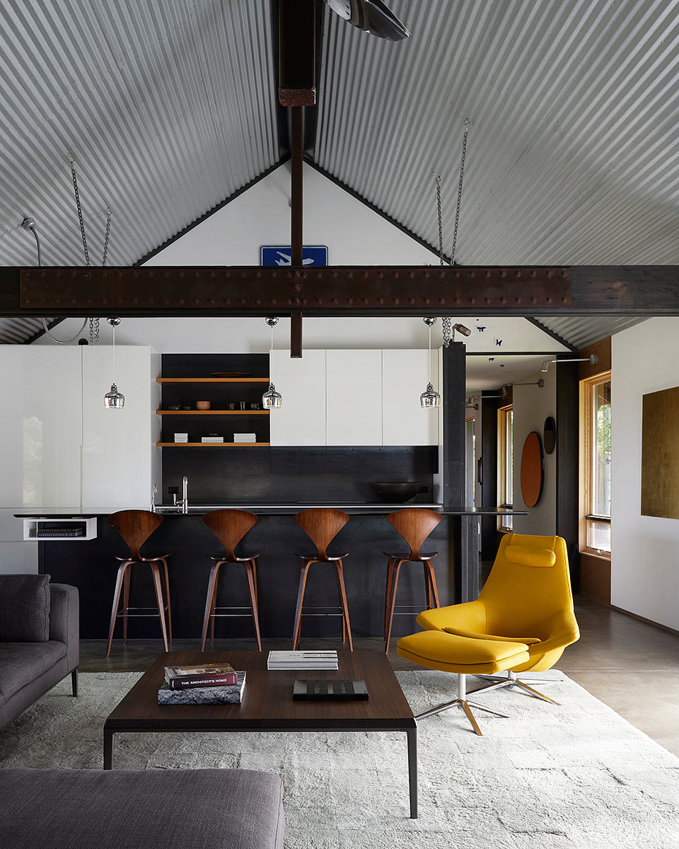 Modern open-concept kitchen featuring sleek black cabinetry, wooden barstools, and a vibrant yellow lounge chair.