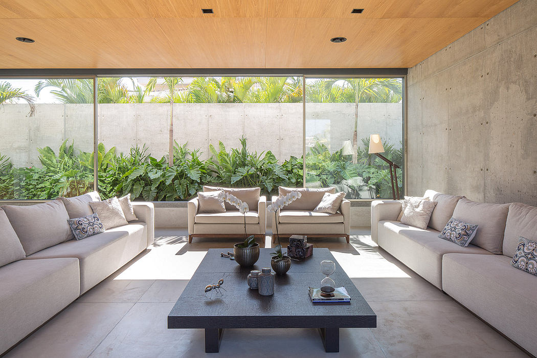Spacious indoor living area with concrete walls, wood ceiling, and large glass windows overlooking lush greenery.