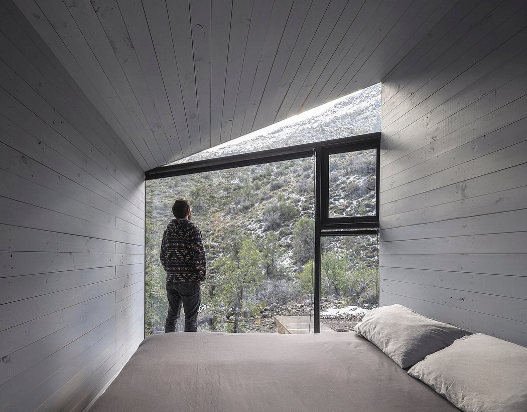 A minimalist bedroom with large window overlooking a snowy landscape, simple furnishings, and wooden paneling.