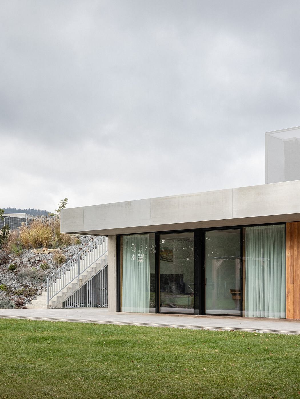 A modern, minimalist building with large glass walls, concrete exterior, and a lush green lawn.