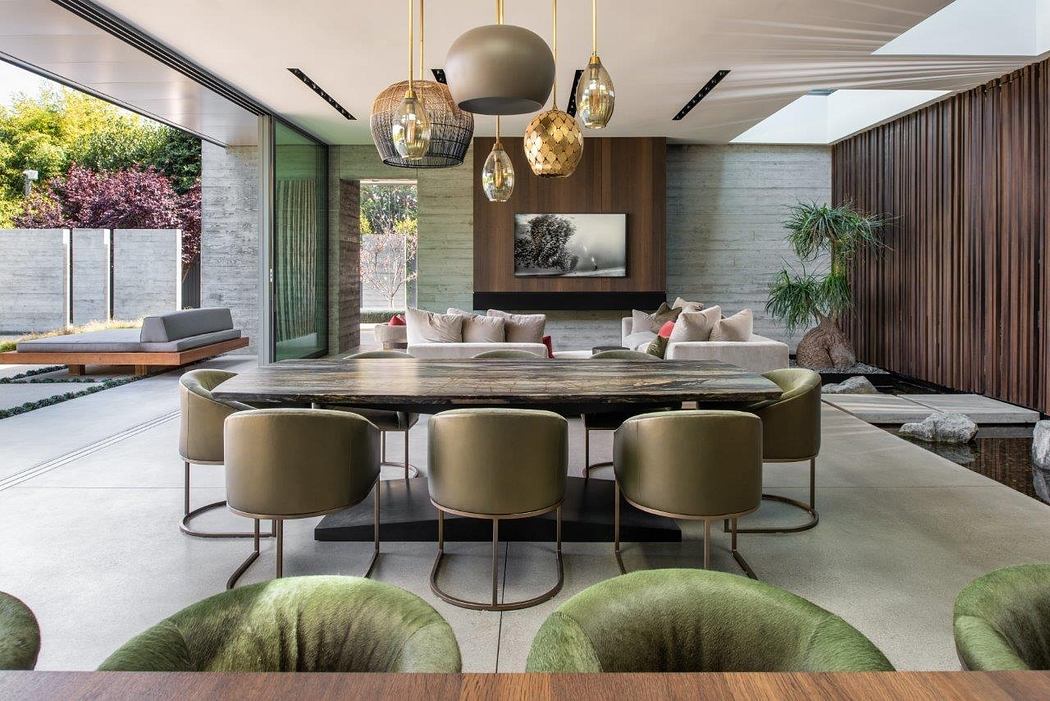 A modern, open-plan living space with a large, stone dining table, plush green seating, and elegant pendant lighting.
