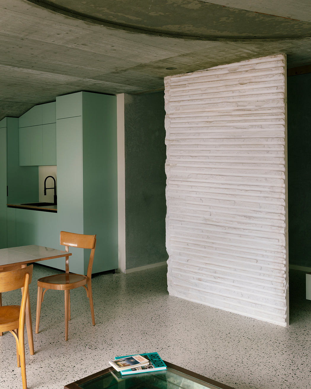 A minimalist kitchen with a white stone wall, green cabinets, and wooden furniture.