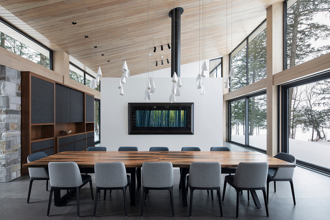 Large modern dining room with wooden ceiling, stone wall, and floor-to-ceiling windows overlooking forest.
