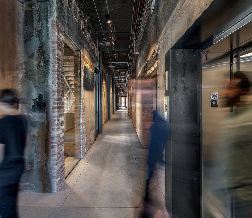 A dimly lit industrial-style hallway with exposed brick walls, concrete floors, and blurred figures moving through.