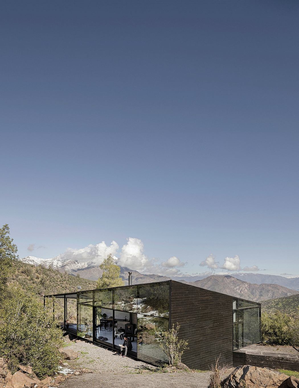 Minimalist cabin nestled in rugged mountainous landscape, with glass walls and metal framework.