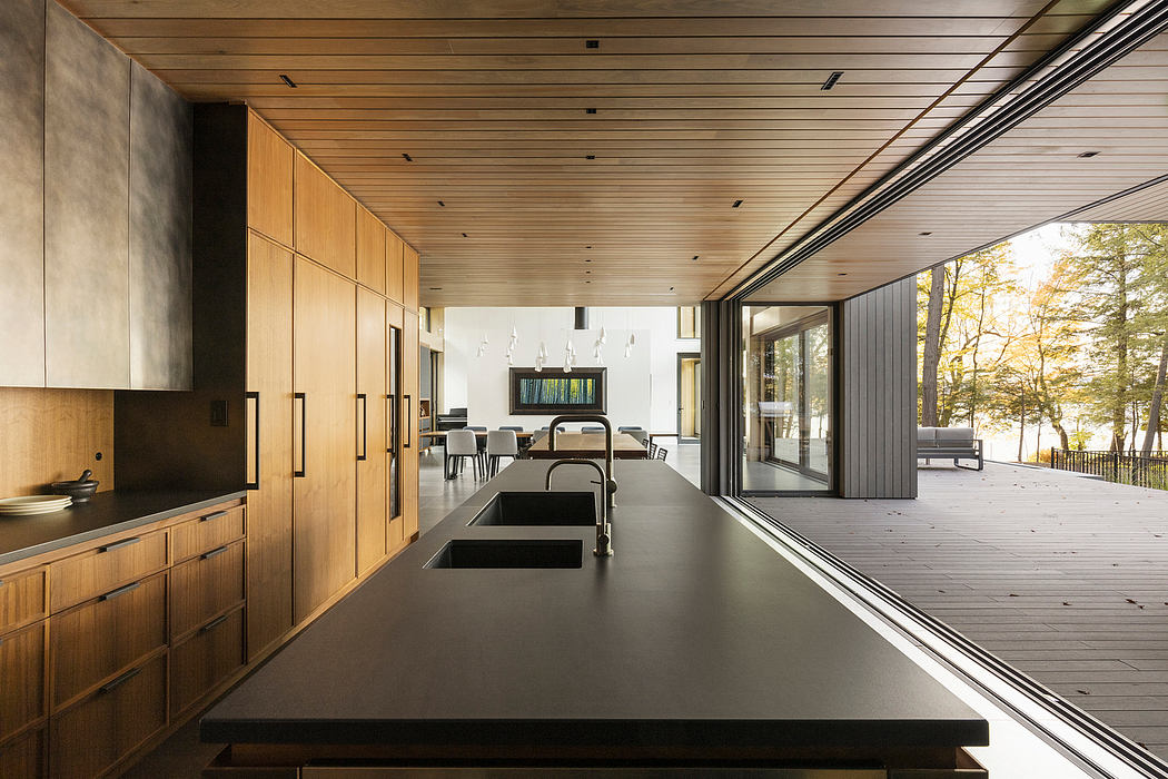 A modern, open-plan kitchen with wooden cabinetry and expansive glass walls overlooking a forest.