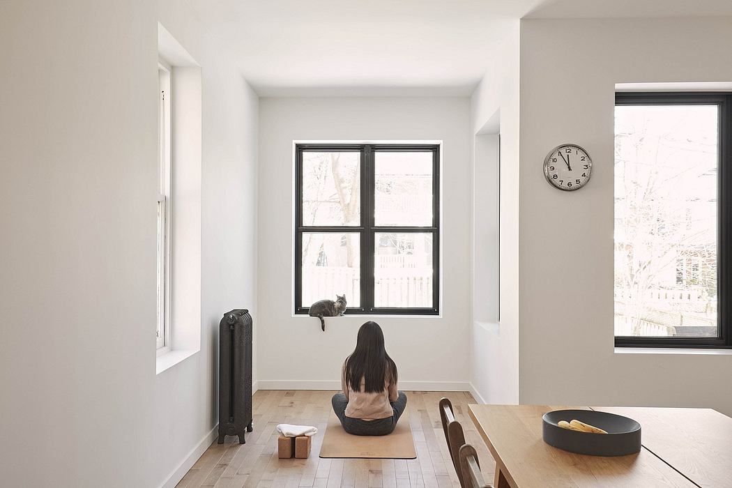 A cozy, minimalist room with large windows, a cat perched on the sill, and a person meditating on the floor.