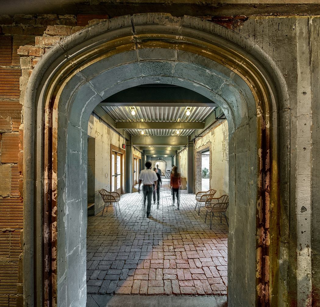 A dimly lit, historic brick archway leads to a long, tiled corridor with simple chairs.