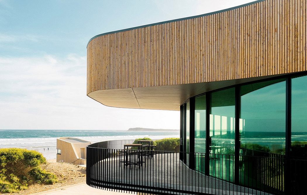 Curved wooden facade overlooking a green landscape and ocean. Glass walls and patio.