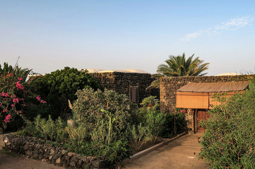 A lush, tropical garden surrounds a rustic stone building with a thatched roof.