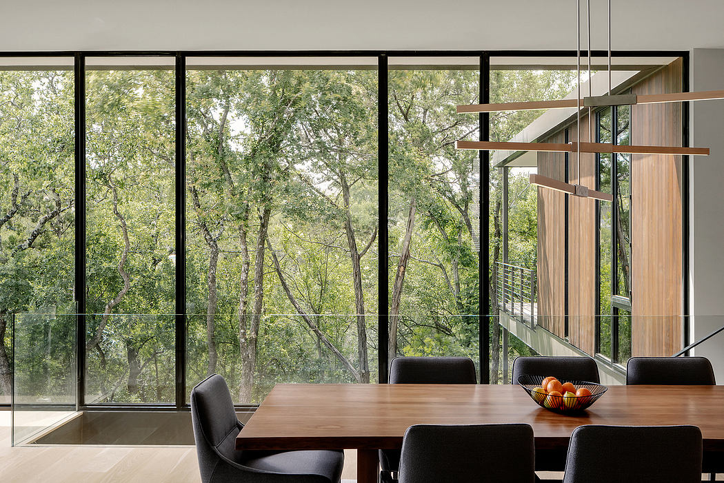Expansive glass walls offer panoramic views of the lush forest surroundings. Wooden dining table and chairs create a warm, modern aesthetic.