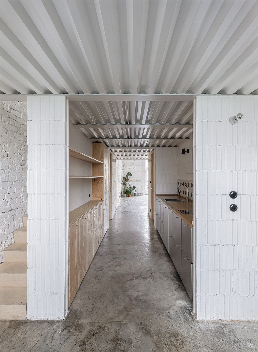 A narrow hallway with white tiled walls and a concrete floor leading to a kitchen with wooden cabinets.