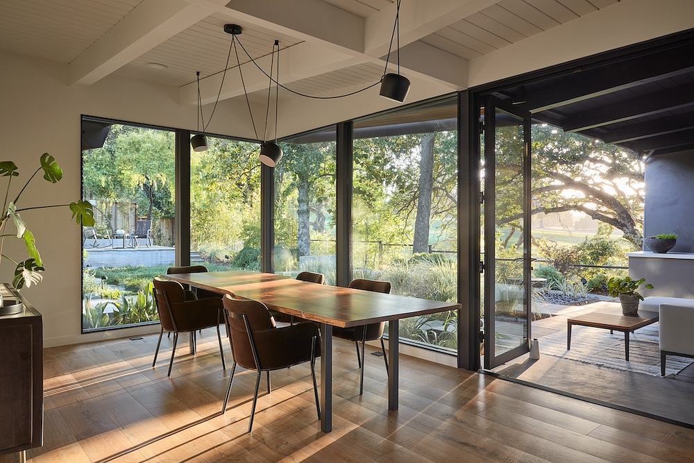 Modern dining room with large windows overlooking lush greenery, wooden table, and pendant lighting.