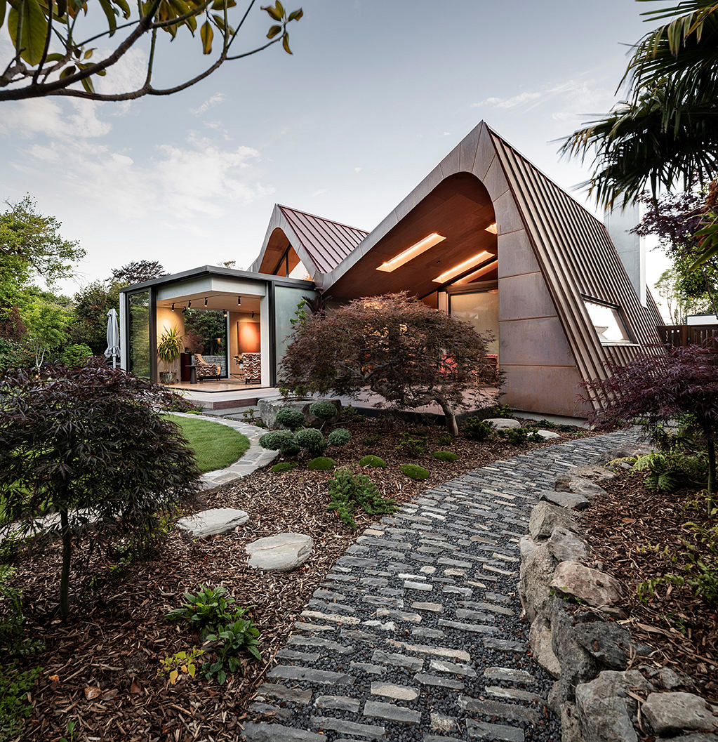 A modern, angular structure with prominent roof lines amid lush greenery and stone path.