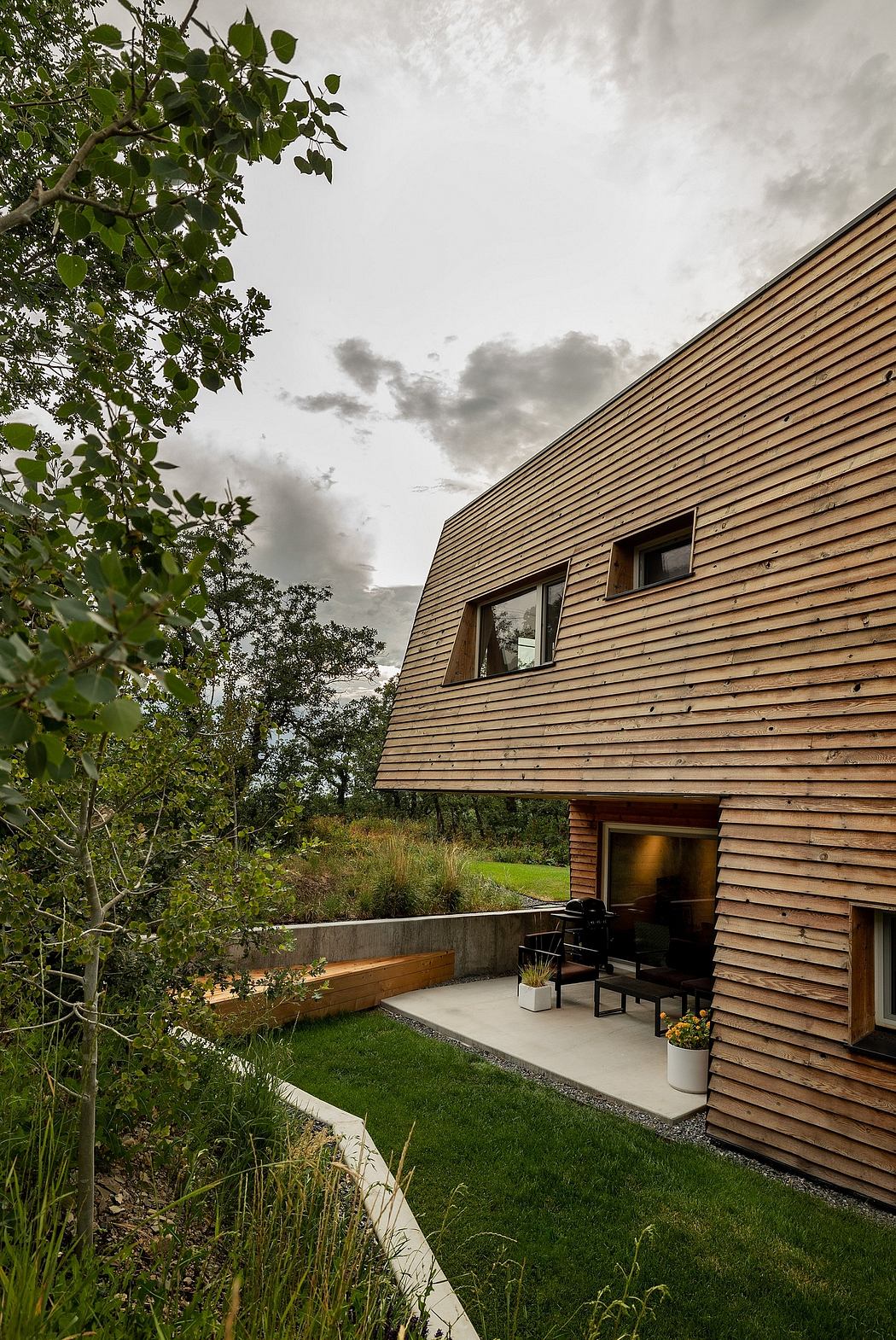 A modern wooden structure with a concrete patio and lush greenery surrounding it.