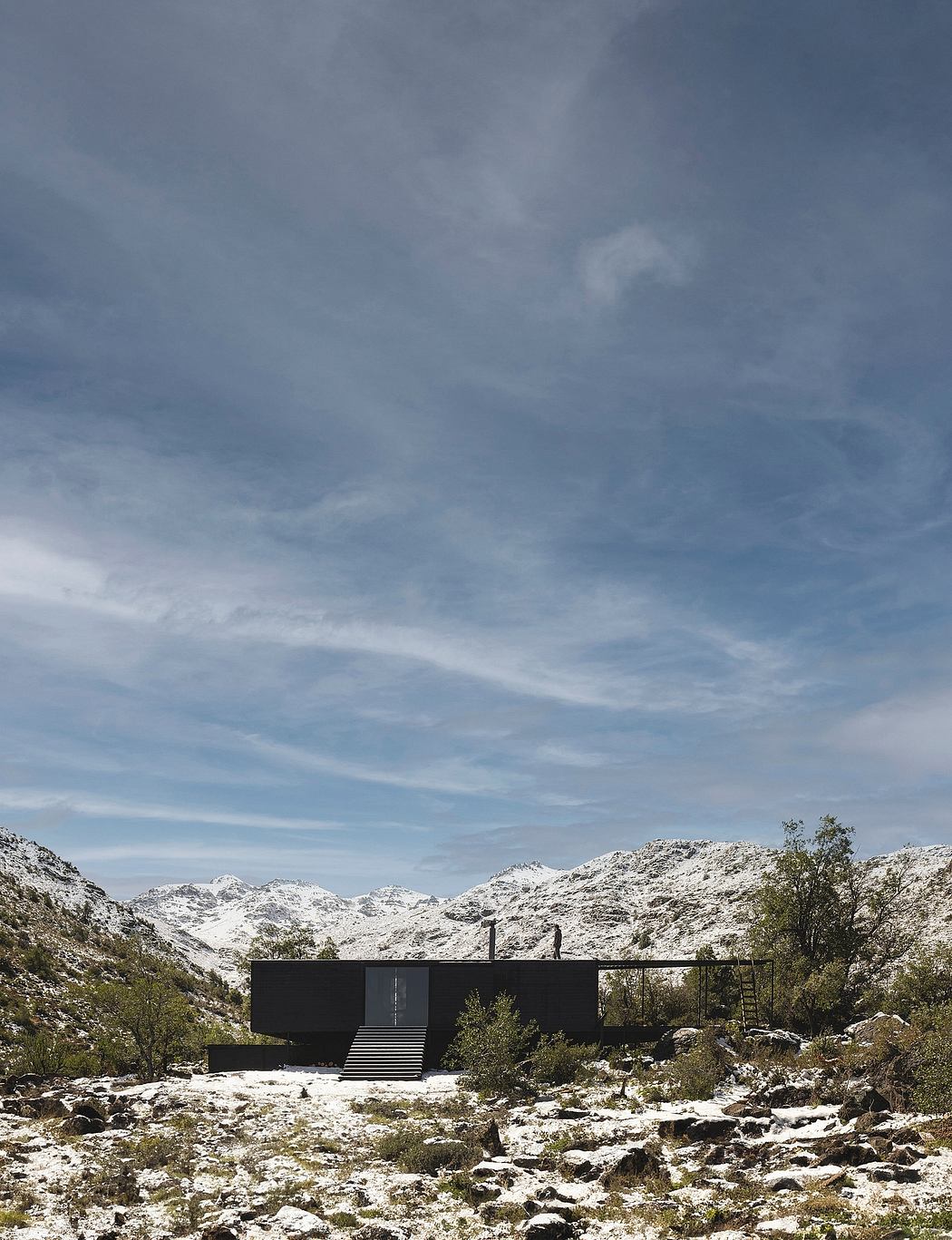 A modern black cabin nestled in a snowy mountainous landscape, with a ladder leading to the rooftop.