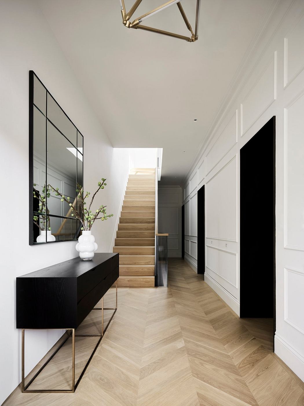 A modern, minimalist foyer with a sleek black console, mirrored panels, and a wooden staircase.