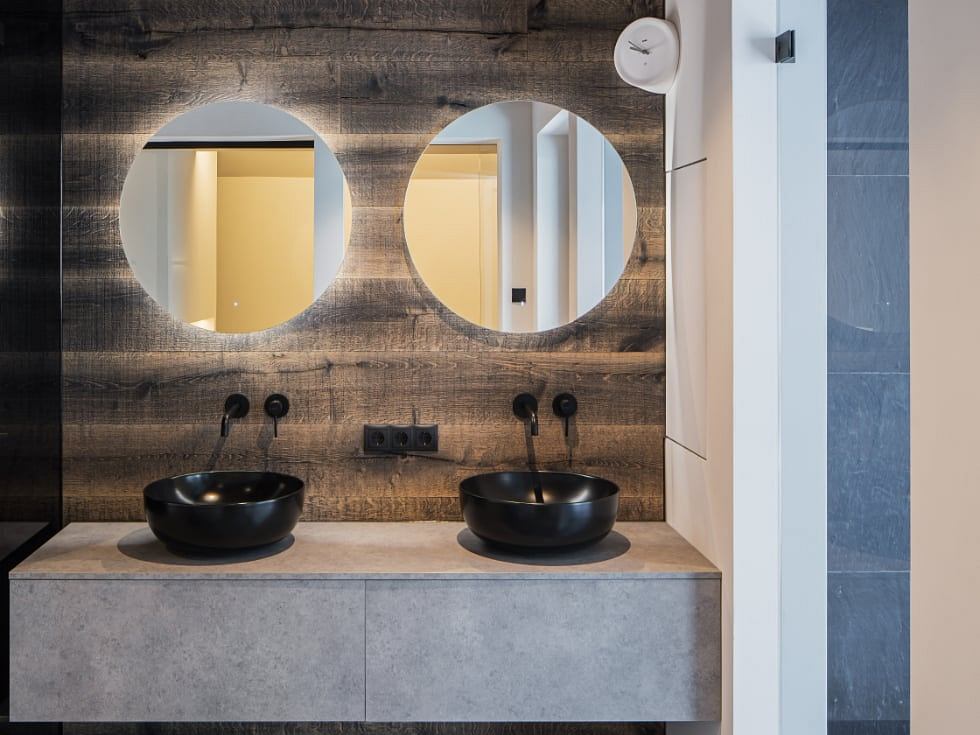 Modern bathroom vanity with round mirrors, black sink bowls, and rustic wood wall.