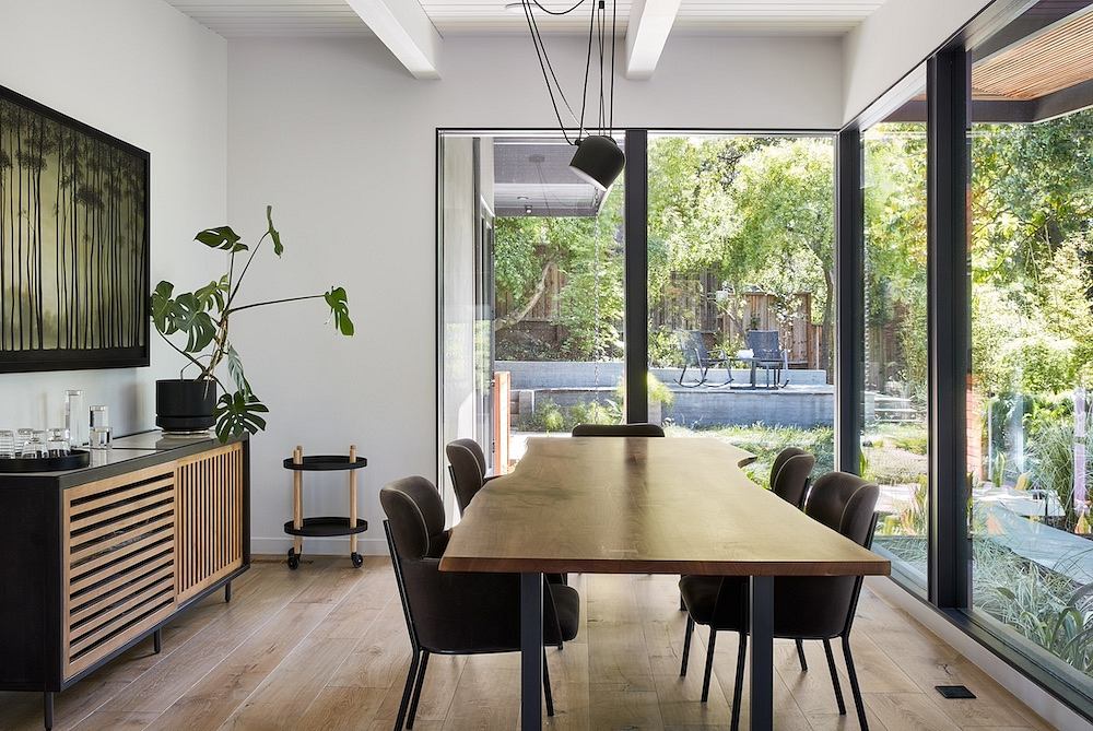 Modern open-concept dining room with wooden table, black chairs, and large windows overlooking lush greenery.