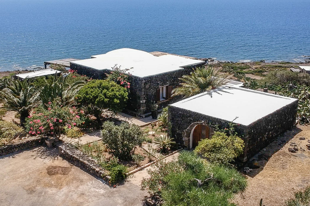 Stone buildings with white roofs and lush surrounding vegetation overlooking the ocean.