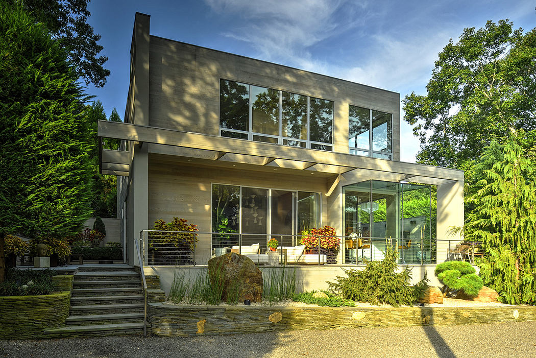 A modern, glass-enclosed house surrounded by lush greenery and a stone patio.