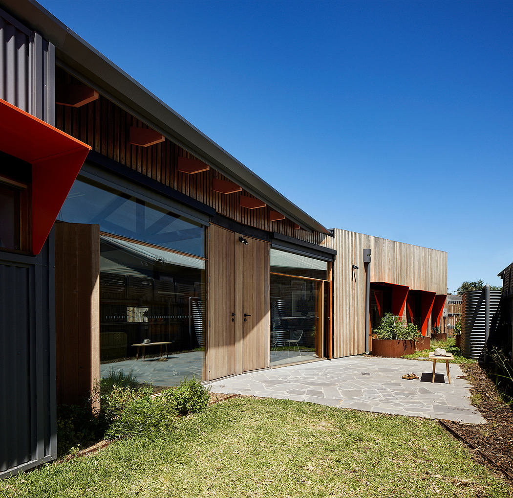 A modern, wooden structure with large glass panels, red accents, and a paved patio area.