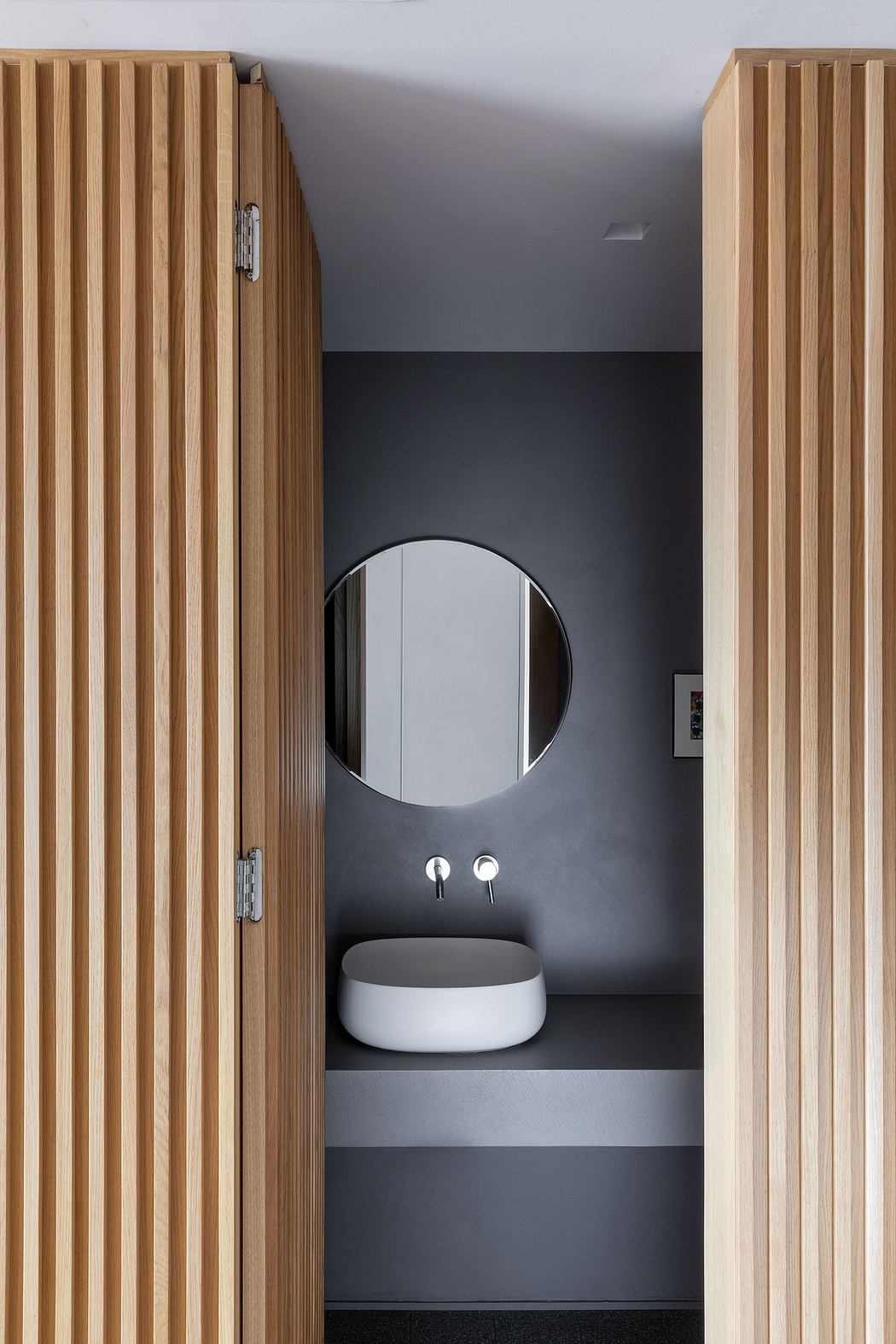 A modern bathroom design with wood paneled walls, a round mirror, and a white vessel sink.