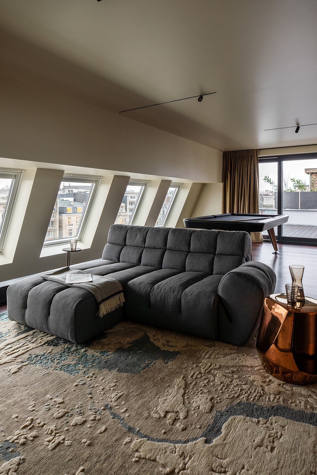 Sleek modern living room with large gray couch, wood-framed windows, and bronze side table.